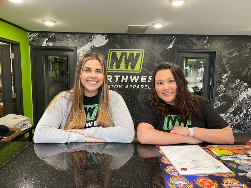 Taylar Hanson and Nika Lao standing behind a counter in the NW Custom showroom. Taylar is late 20's and Nika is in her early 30's. Taylar is wearing  a gray sweater and Nika has on a black tshirt